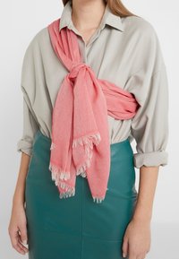 Femme portant une chemise beige à manches retroussées, une jupe en cuir bleu sarcelle et une écharpe corail à franges nouée en un nœud lâche sur la poitrine.