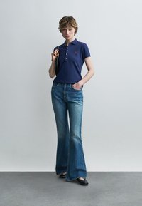 Kvinne iført en marineblå polotrøye og vide blå jeans, holder en svart håndveske over skulderen, stående mot en ensfarget grå vegg.