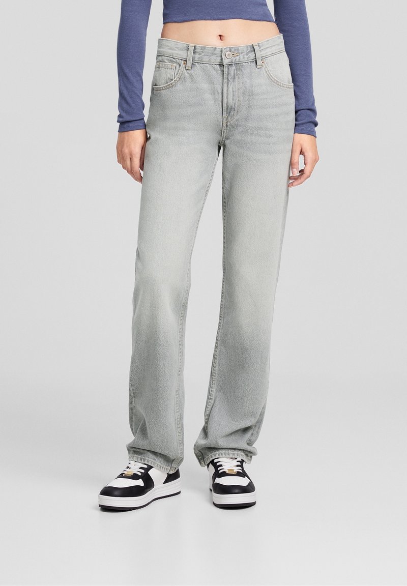 Vaqueros de mezclilla gris claro con un ajuste relajado, diseño de cinco bolsillos y textura desgastada, combinados con zapatillas negras y blancas.
