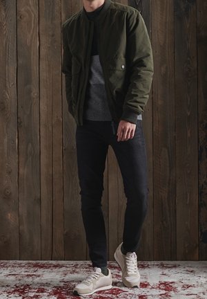 Mężczyzna w ciemnozielonej kurtce, szarym swetrze, czarnych spodniach i białych trampkach stojący przy drewnianej ścianie na czerwono-białym dywanie.