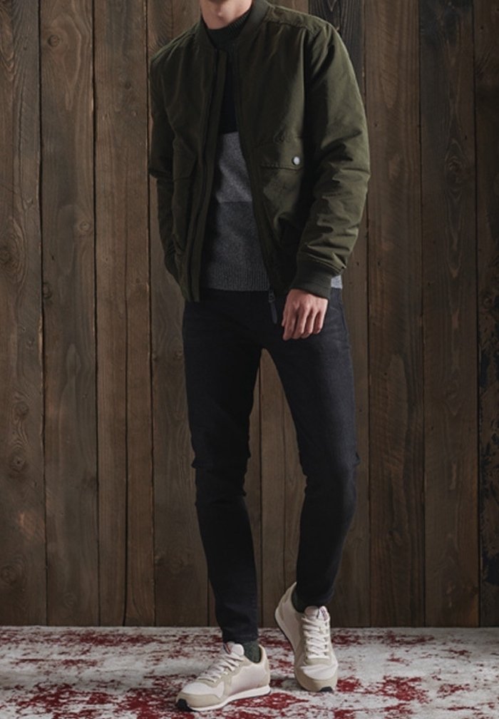 Homme portant une veste vert foncé, un pull gris, un pantalon noir et des baskets blanches, debout contre un mur en bois sur un tapis rouge et blanc.