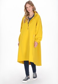 Hosszú sárga esőkabát cipzárral, rugalmas mandzsettákkal és kapucnival, csíkos felsővel, sötét farmerrel és világos sportcipővel párosítva.