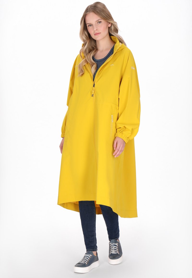 Hosszú sárga esőkabát cipzárral, rugalmas mandzsettákkal és kapucnival, csíkos felsővel, sötét farmerrel és világos sportcipővel párosítva.