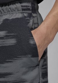 Šedé sportovní šortky s texturovaným, abstraktním tmavým vzorem, s elastickým pasem a bočními kapsami. Hladký povrch tkaniny.