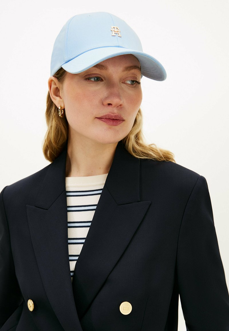 Mujer con gorra azul claro con logo dorado, pendientes de aro dorados, blazer azul marino cruzado con botones dorados y camisa a rayas debajo.