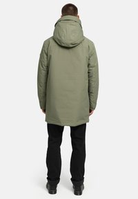 Olivno zelena vodoodporna parka s kapuco, dolga, iz teksturiranega materiala in z zatesnjenimi šivi. Nosi se over črnimi hlačami in trdno obutvijo.
