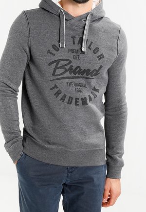 Homme portant un sweat à capuche gris avec le texte "Tom Tailor Brand" et un pantalon bleu marine, les mains partiellement dans les poches, debout devant un arrière-plan uni.