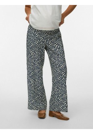 Persona che indossa pantaloni a taglio largo con motivi a foglia blu scuro e scarpe marroni con tacco basso, in piedi contro uno sfondo neutro.