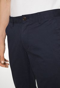 Tmavě modré kalhoty vyrobené z bavlněné směsi, s knoflíkovým zapínáním, poutky na opasek a bočními kapsami s hladkým povrchem.