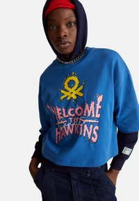 Oříznutá modrá mikina s potiskem "Vítejte v Hawkins" v růžové barvě, se žlutými detaily a černou kapucí. Obsahuje řetízkový detail a nášivku s logem.