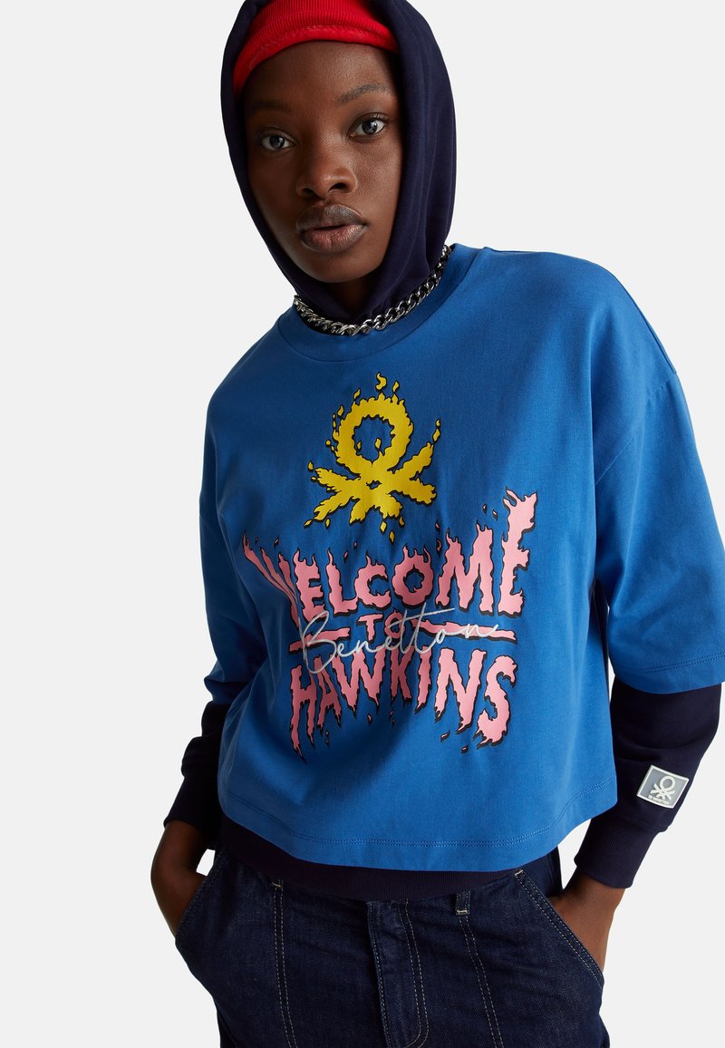 Oříznutá modrá mikina s potiskem "Vítejte v Hawkins" v růžové barvě, se žlutými detaily a černou kapucí. Obsahuje řetízkový detail a nášivku s logem.