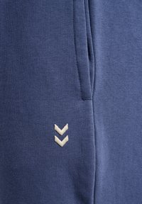 Blauwe katoenen joggers met een zachte textuur, ribbelstof en een crèmekleurig geborduurd logo met een chevrondesign aan de linkerkant.