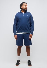 Άνδρας με πλεξούδες, φορώντας μπλε σκούρο φούτερ με φερμουάρ μέχρι τη μέση, ταιριαστά σορτς, λευκές κάλτσες και μαύρα αθλητικά παπούτσια, στέκεται μπροστά σε απλό φόντο.