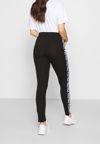 Žena v černých legínách s bílými pruhy s logem Fila po stranách a bílých teniskách, stojící na jednobarevném pozadí.