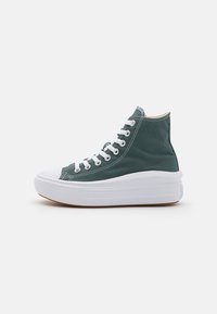 Adidași cu vârf înalt, din pânză verde, cu șireturi albe, vârf proeminent și o talpă groasă albă, cu un fond texturat maro.