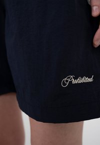 Gros plan sur la jambe d'une personne portant un short sombre brodé du mot « Interdit » en cursive près de l'ourlet.