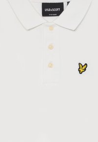 Polo shirt blanc pour enfants avec trois boutons, logo d'aigle jaune sur la poitrine et étiquette de marque Lyle & Scott pour 9-10 ans.