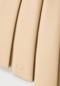 Μπεζ πλισέ ύφασμα με μικρό ανάγλυφο λογότυπο κροκόδειλου κοντά στην κάτω άκρη σε λεία επιφάνεια.