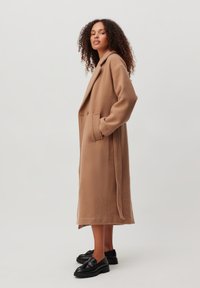 Lange beige jas gemaakt van zachte, gestructureerde stof, met een dubbelzijdig ontwerp, zijzakken en een lengte tot halverwege de kuit. Draagt met zwarte schoenen.
