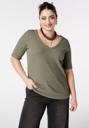 Vrouw die glimlacht en een olijfgroene V-hals top met korte mouwen draagt, zwarte spijkerbroek en gelaagde kettingen met gouden oorbellen tegen een witte achtergrond.