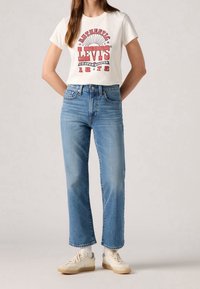 T-shirt blanc en coton avec texte graphique rouge, associé à un jean bleu clair droit et des baskets beige. La tenue présente un style décontracté.