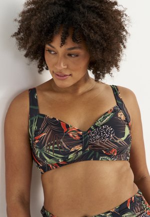 Kobieta z kręconymi włosami, ubrana w górę bikini z tropikalnym kwiatowym wzorem, patrząca w dół i w prawo na jednolitym jasnym tle.