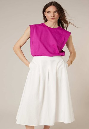 Femme aux longs cheveux bruns portant un haut sans manches rose vif et une jupe midi blanche taille haute, mains dans les poches, fond neutre.