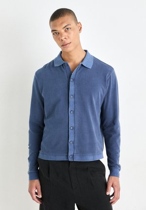 Blue long-sleeve shirt with a button-up front, contrasting light blue collar, smooth texture, and a relaxed fit. Styled with dark trousers.