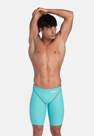 Sportlicher Mann dehnt sich mit über dem Kopf ausgestreckten Armen, trägt türkisblaue Arena Schwimmjammers vor schlichtem hellem Hintergrund.