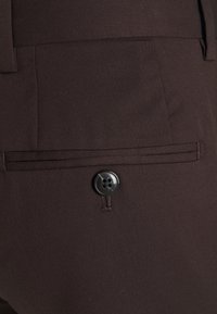 Bruna tygbyxor med en slät yta, med knappstängning och sydda detaljer. Inkluderar en bakficka med en enkel knapp.