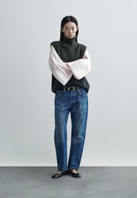 Pull à col roulé vert épais et blouse rose clair avec manches plissées, associée à un jean bleu droit et des mocassins noirs.
