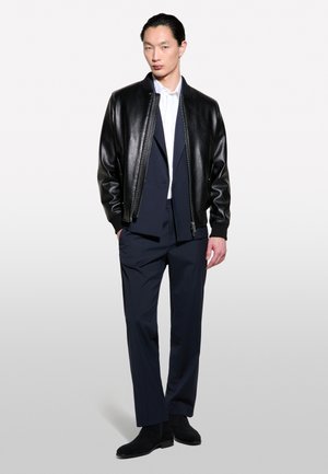 Homme portant une veste en cuir noire sur un costume bleu marine et une chemise blanche, debout devant un fond clair uni.