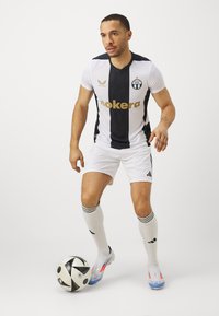 Maglia da calcio a righe bianche e nere con scritte dorate, abbinata a pantaloni corti e calze bianche. Il giocatore è in azione con una palla.