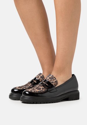 Mocassins en cuir verni noir avec des semelles épaisses et un dessus en imprimé léopard portés pieds nus sur fond blanc.