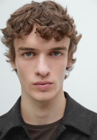 Portret młodego mężczyzny o kręconych, jasnobrązowych włosach, ubranego w ciemną koszulę na guziki, nałożoną na brązowy top. Jego oczy są jasnozielone, a wyraz twarzy neutralny.