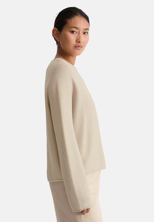 Pull en tricot beige avec une coupe décontractée, un col rond et des manches longues. Le tissu est texturé avec un motif subtil.