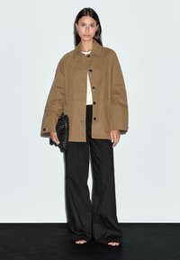 Bruine oversized jas met zwarte knopen en zakken, gecombineerd met zwarte wijde broek. Accessoires zijn een gestructureerde zwarte clutch.