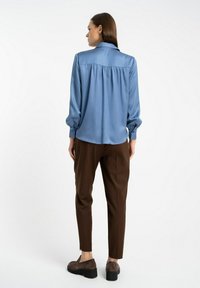 Jasnoniebieska bluzka z długim rękawem i kołnierzykiem, z marszczonym tyłem, w zestawie z brązowymi rurkami i brązowymi butami na obcasie. Gładka faktura materiału.