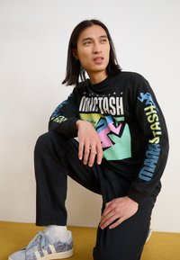 Black long-sleeve shirt with colorful graphic design and text, paired with dark pants and light blue sneakers featuring a patterned accent.