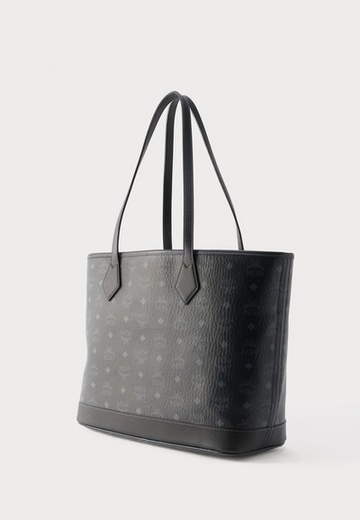 Sac fourre-tout en cuir noir texturé avec de longues poignées et un motif subtilement embossé, posé sur un fond clair uni.