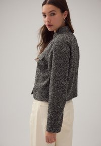 Teksturowana czarno-biała tweedowa kurtka o krótkim kroju, z zaostrzonym kołnierzem i długimi rękawami, zestawiona z jasnymi spodniami.