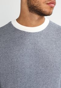 Granatowy sweter dziany z teksturowanym wzorem, z białym, prążkowanym, okrągłym dekoltem. Zbliżenie na tors i obszar szyi.