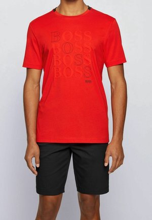 Mężczyzna w jasnoczerwonej koszulce z krótkim rękawem z powtarzającym się napisem „BOSS” oraz czarnych, jednolitych szortach do kolan, stojący na jasnym tle.