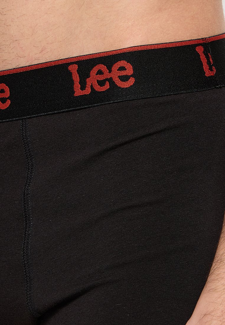 Gros plan sur la ceinture de sous-vêtements Lee noirs avec du texte rouge, située au niveau de la taille et de la hanche d'une personne.
