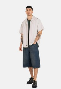 Hellgraues kurzärmliges Hemd, grünes Unterhemd, Jeansshorts, schwarze Schuhe mit metallischen Akzenten und sichtbare Tattoos an den Armen.