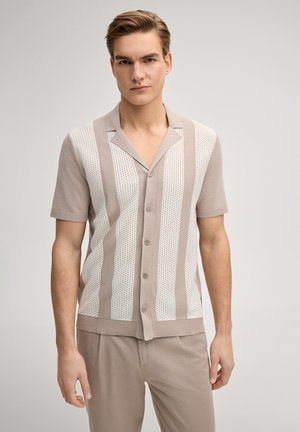 Junger Mann, der ein beige gestricktes, kurzärmeliges Hemd mit senkrechten weißen Lochstreifen und passende beige Hosen trägt, steht neutral da.