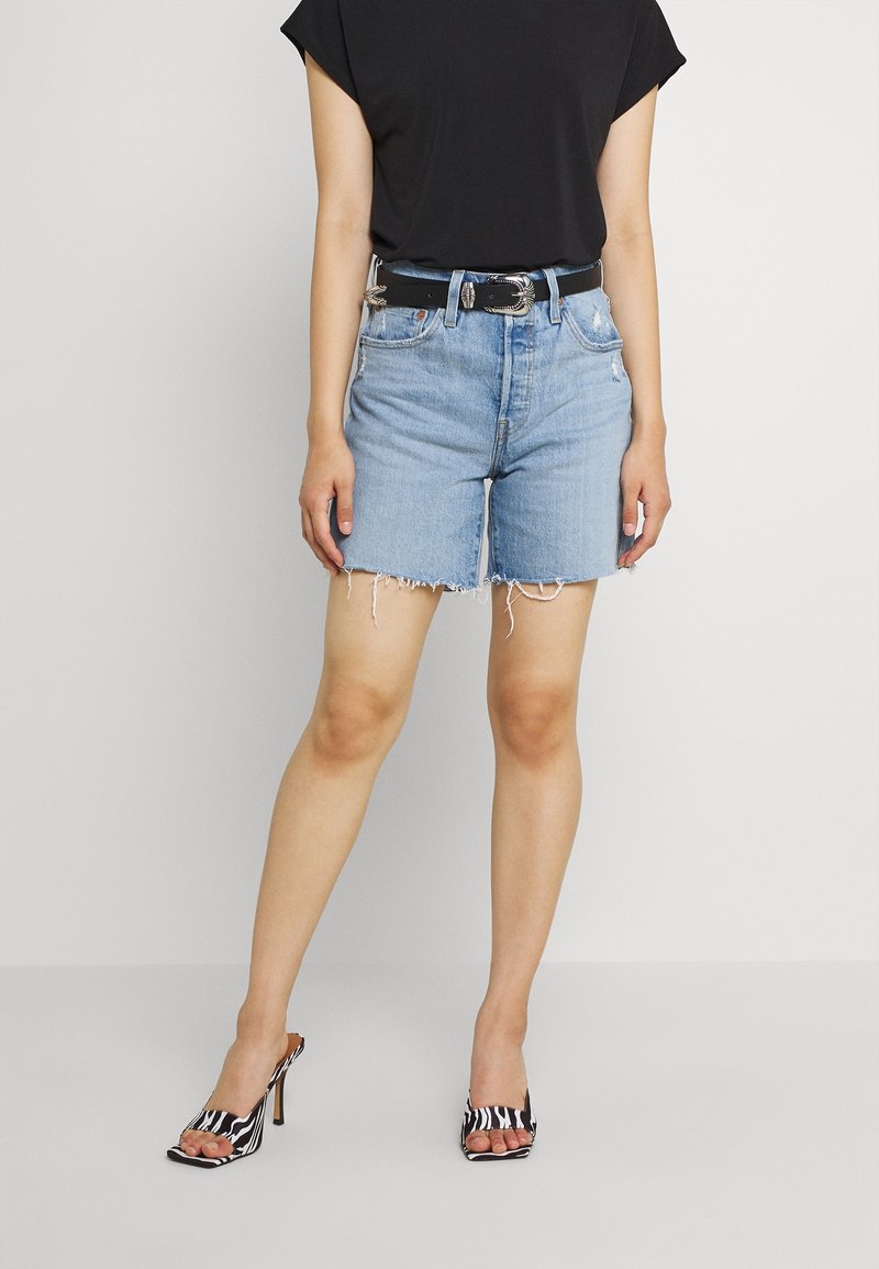 Mujer con shorts de mezclilla desgastados color azul claro, cinturón negro, camiseta negra metida por dentro y sandalias de tacón con estampado de cebra en blanco y negro.