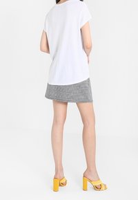 Mujer con camiseta blanca de manga corta, minifalda de cuadros gris y sandalias amarillas de tacón alto, de pie con la espalda hacia adelante.