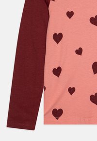 Langarmshirt met een lichtroze lichaam en kastanjebruine mouwen; heeft een patroon van donkerrode harten aan de voorkant. Gemaakt van katoen.
