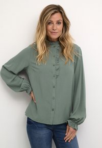 Blusa verde de manga larga con detalles de volantes a lo largo del placket de botones. Tejido suave, puños ajustados y un brillo sutil, combinada con jeans azules.